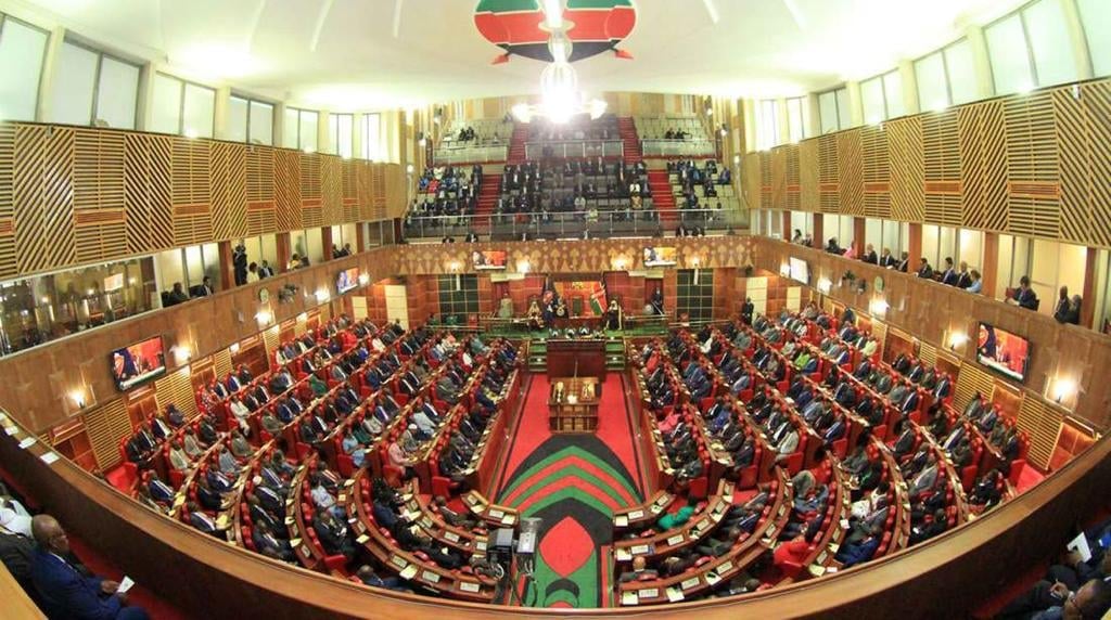 Interior of a legislative parliament chamber with red seating, ornate wooden walls, and galleries filled with members during a session