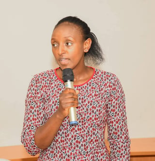 Woman in a burgundy floral top speaking into a microphone, standing against a beige background
