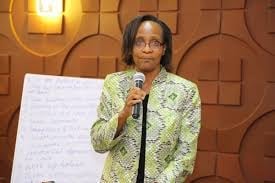 Woman in green patterned jacket speaking at microphone with whiteboard behind her