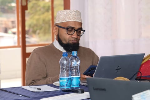 Bearded man wearing glasses and a white cap, seated at a desk with a laptop, water bottle, and notepad
