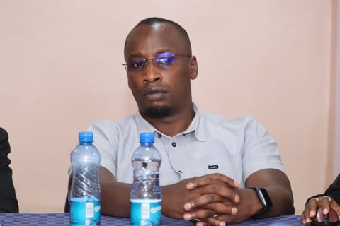 Man wearing light blue shirt and blue-tinted glasses sits at table with water bottles in front of him during what appears to be a panel or meeting
