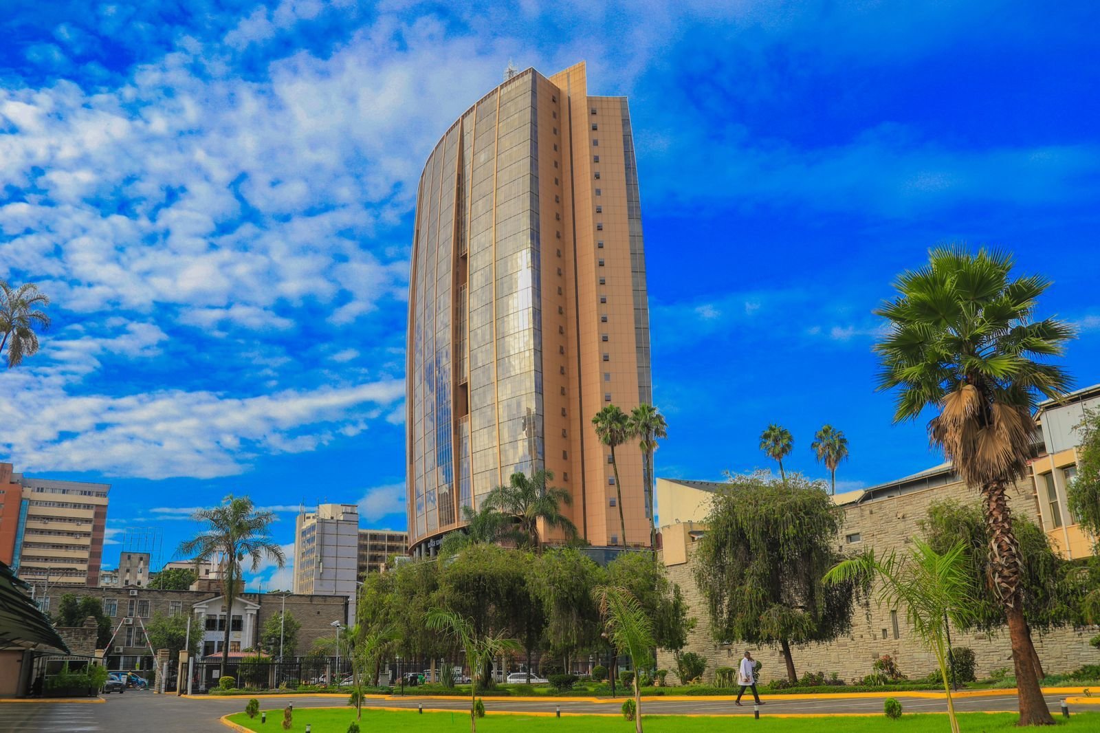 Tall brick high-rise building surrounded by palm trees and green lawn under a bright blue sky with clouds
