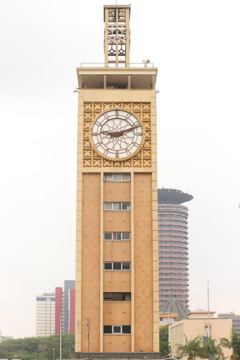 Tall brick clock tower with ornate geometric patterns and clock face, surrounded by modern buildings in urban skyline