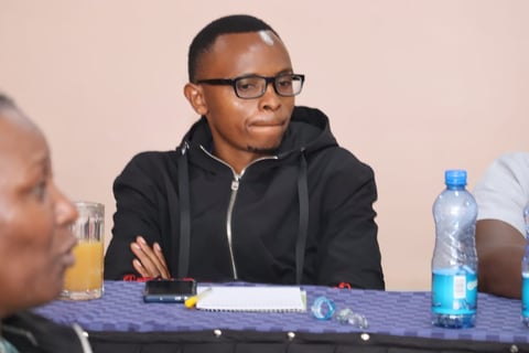 Man wearing black hoodie and glasses sits at conference table with water bottle and notebook