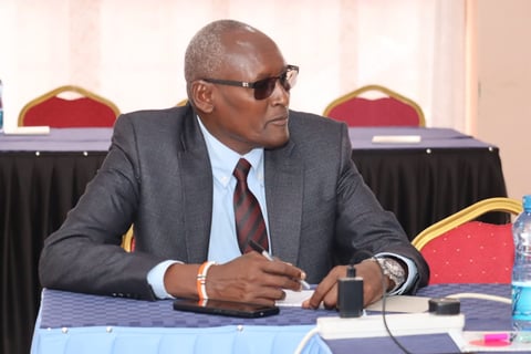Older man in gray suit and sunglasses sitting at conference table with microphone during formal meeting