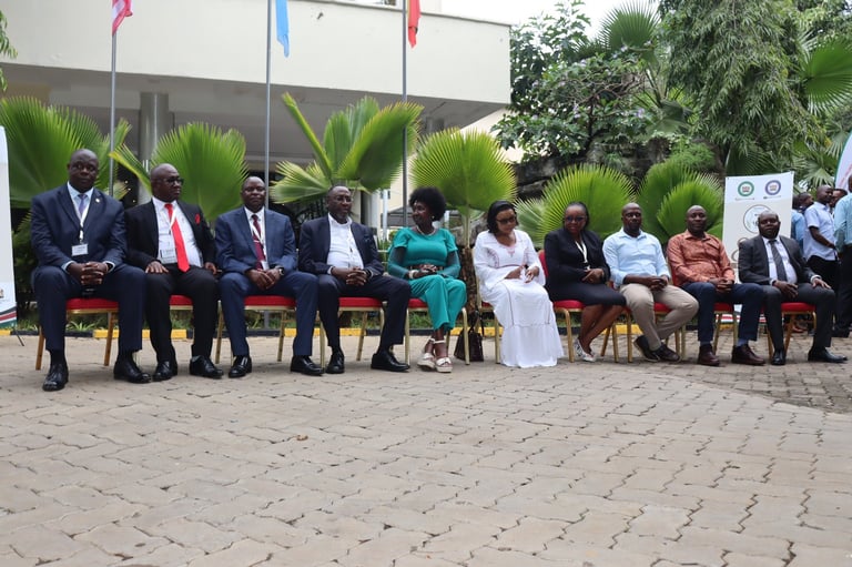 Group of formally dressed people seated outdoors on red chairs, with flags and tropical plants in background